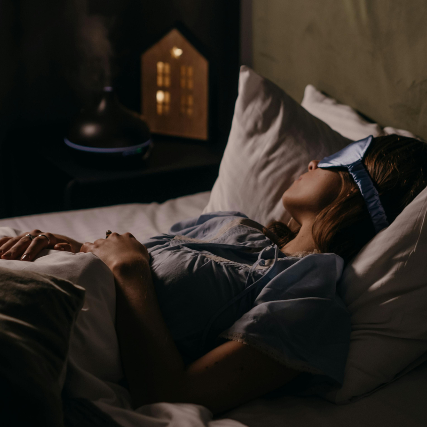 A woman wearing a satin sleep mask lies on a pillow in a bed.