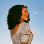 A black woman stands in front of the bright night sky, the Full Moon seen in the background.