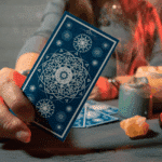 Someone (not pictured) holds up a blue Tarot card with crystals and a candle in the background.