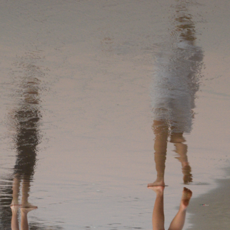 Two people stand next to water, their reflections primarily showing.