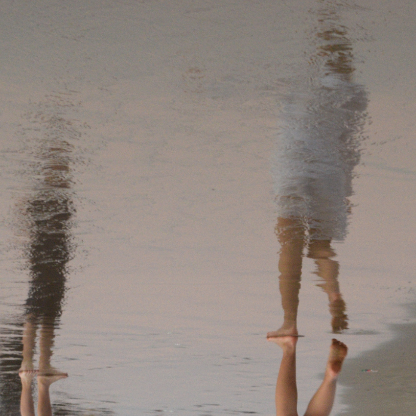 Two people stand next to water, their reflections primarily showing.