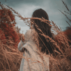 A brunette woman wears a cozy-looking cream sweater. She is facing away from the camera and there are autumnal looking wheat stalks around her.