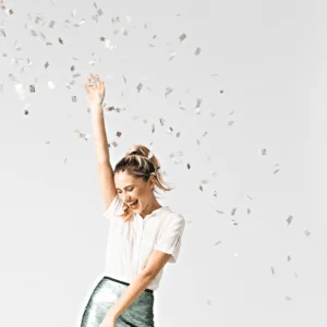 blonde woman in a white shirt and green skirt dancing gleefully with confetti in the air
