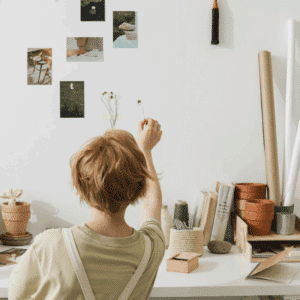 A person with short, red hair holds up a small dried flower against a wall with art supplies and other photos.