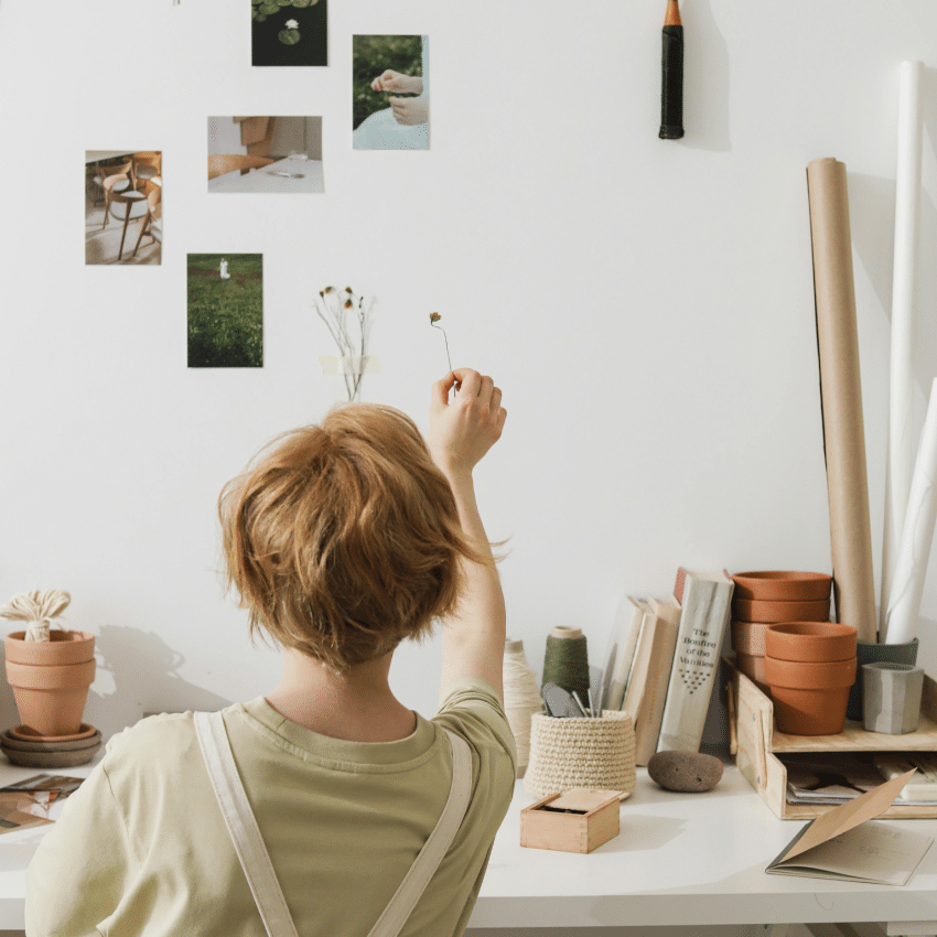 A person with short, red hair holds up a small dried flower against a wall with art supplies and other photos.