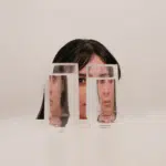 A woman's face appears behind 2 glasses of water, the water distorts her image representing the energy of Jupiter retrograde in Cancer.