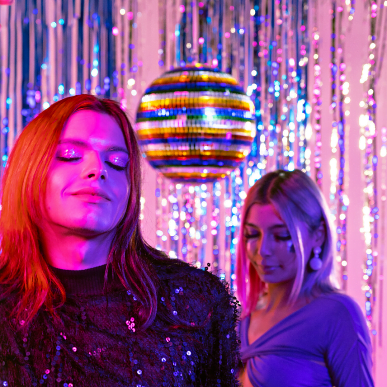 two women in sparkly outfits standing underneath a disco ball against a glittery backdrop