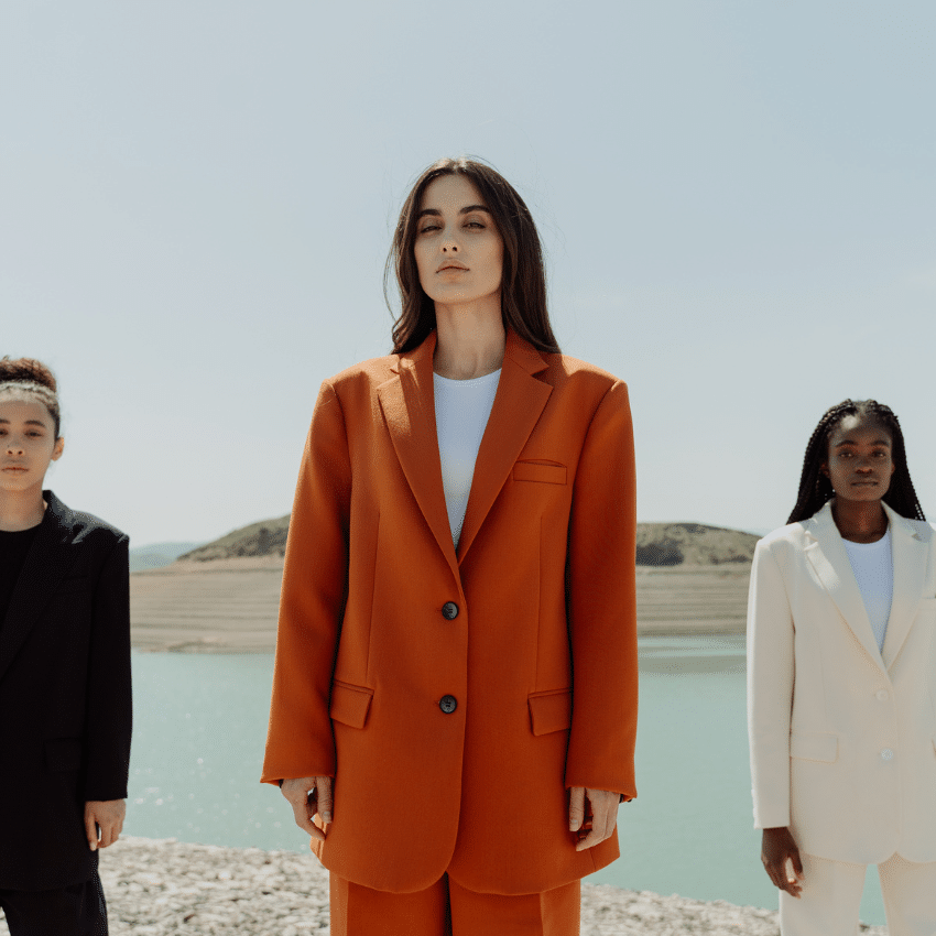 brunette woman in an orange suit standing in front of a lake with two other women in suits on either side of her