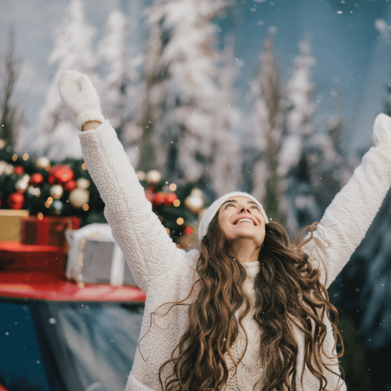 gleeful woman with long brown hair with her hands in the air smiling as snow falls around her with Christmas presents in the background