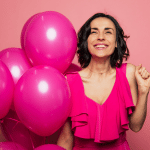 gleeful woman in pink with short brown hair smiling holding pink balloons