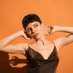 woman with short brown hair in a sparkly brown dress posing with her arms in the air against an orange backdrop