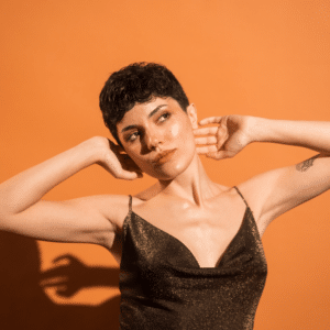 woman with short brown hair in a sparkly brown dress posing with her arms in the air against an orange backdrop