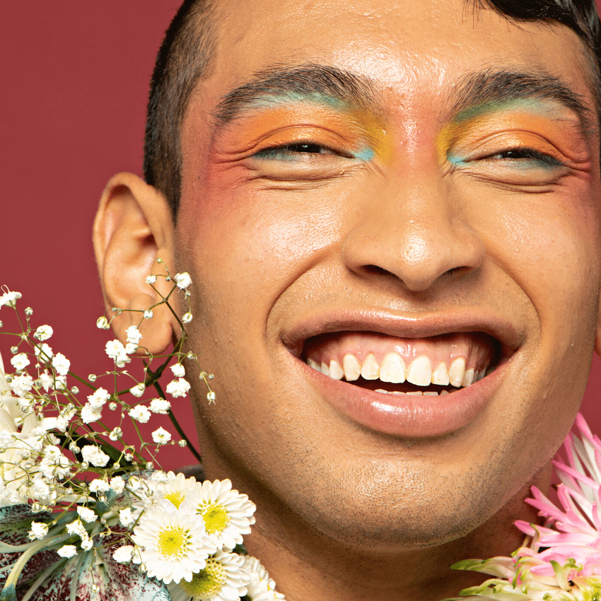 man with colourful eyeshadow smiling boldly with flowers around his face