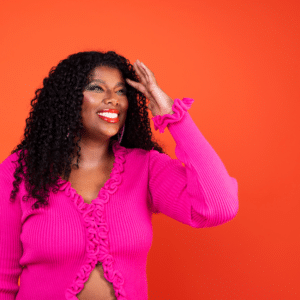 woman in bright pink shirt and funky eyeshadow staring ahead against a bright orange backdrop