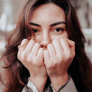 A woman with dark brown hair holds her hands in fists up to her mouth, as if she has a secret to reveal.