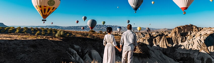 Two figures stand with their backs visible, holding hands. They are looking at many hot air balloons floating in the sky.