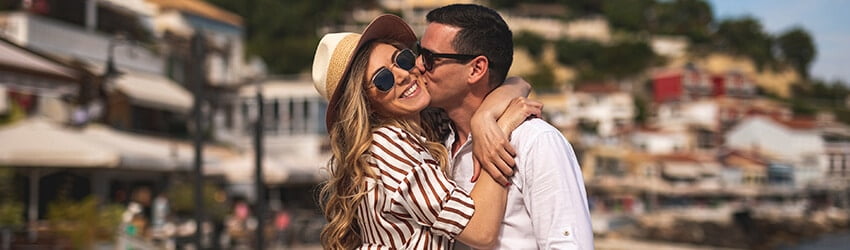 A man and a woman wearing sunglasses embrace. The man kisses the woman on the cheek.