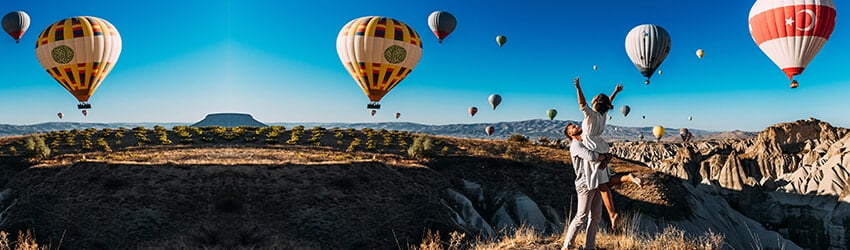 Two figures reach their arms into the air as if to catch the hot air balloons in the air behind them.