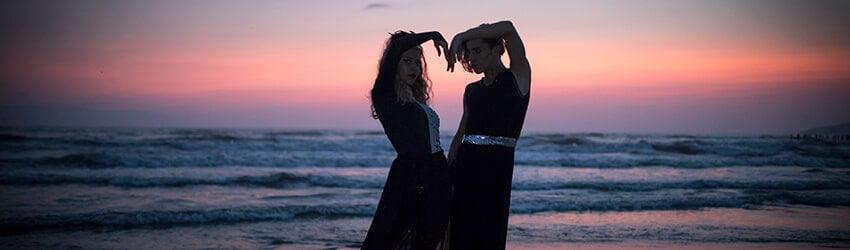 Two figures use their arms to form a heart. They stand near the beach at dusk.
