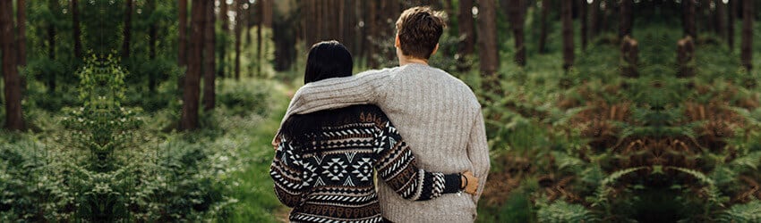 Two figures stand in the forest with their arms around each other.