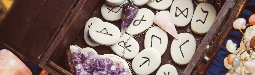 Cream-colored rune stones fill a brown wooden box that also contains a piece of raw amethyst, an amethyst crystal pendulum, and a rose quartz pendulum.