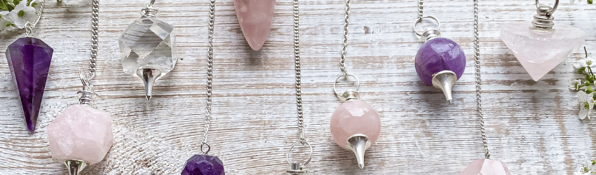 Different shapes and sizes of pink, purple, and clear crystal pendulums lay on a wooden table.