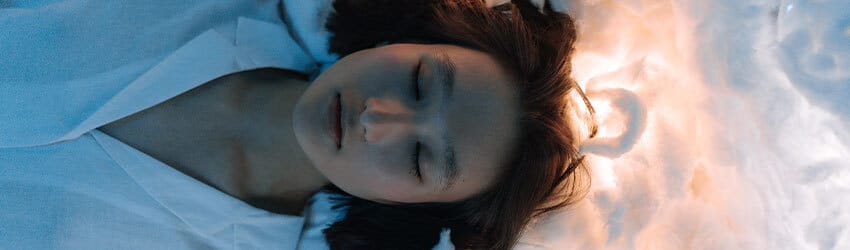 A woman sleeps with her head on a white pillow and the space behind her head is illuminated.