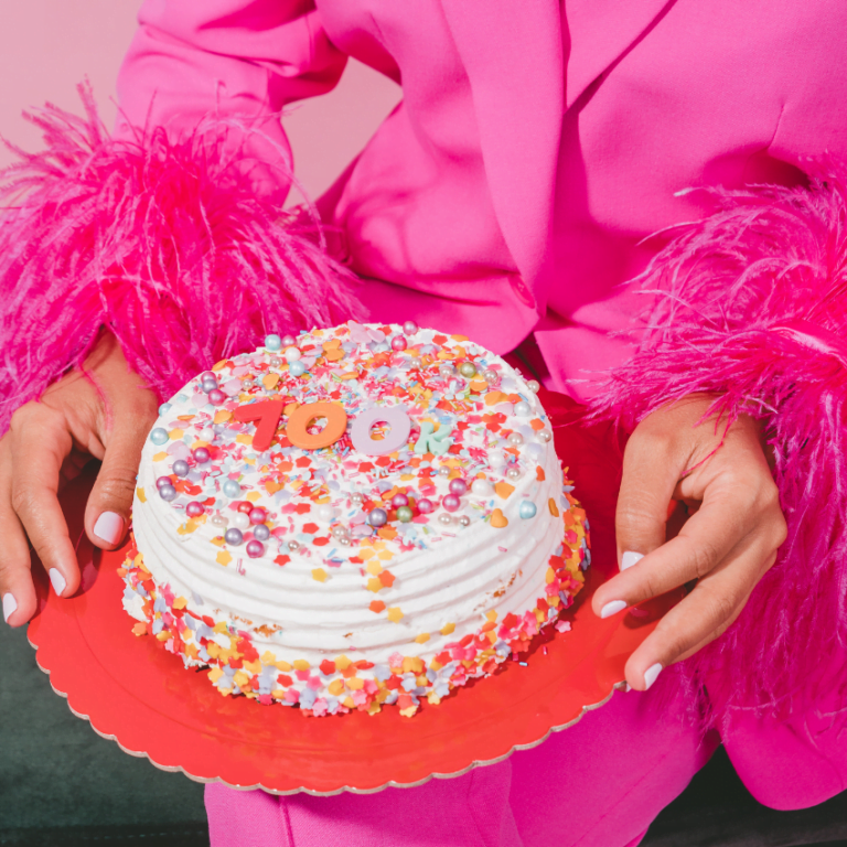 woman in a pink suit holding out a brightly decorated cake