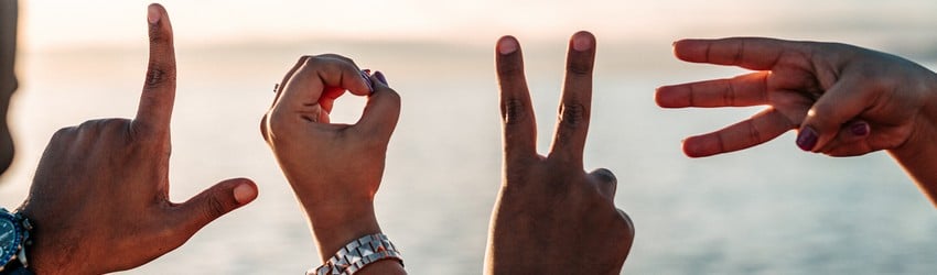 People hold their hands up to spell LOVE with their fingers on a beach at sunset.