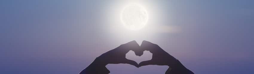 A woman holds her hands up in front of the Full Moon in a heart shape.