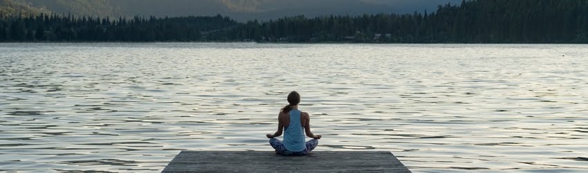 A woman sits in a meditative cross-legged pose at the end of a wooden dock. She is facing a large body of water with mountains in the background. It is a dreary, yet peaceful scene.