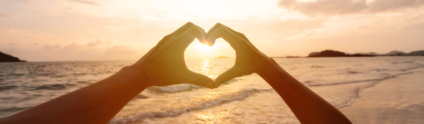 A man and woman's hands form a heart with the ocean and sunset in the background.