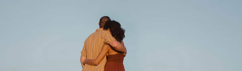 A couple holds each other on a beach at sunset while looking at the distant full moon rising.