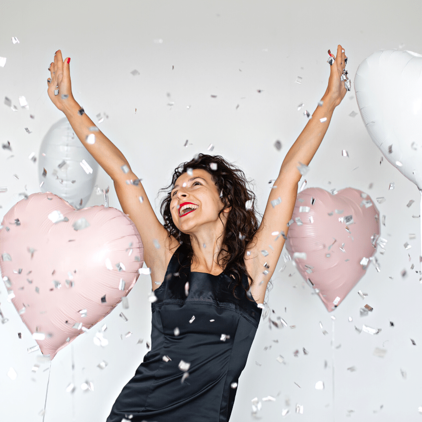 gleeful brunette woman in a black dress and red lipstick with her hands in the air surrounded by heart-shaped balloons and confetti