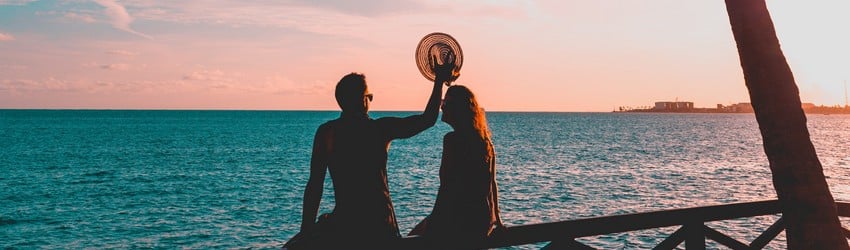 Two people sit on a railing near the ocean watching the sunset. They are falling in love in the summer.