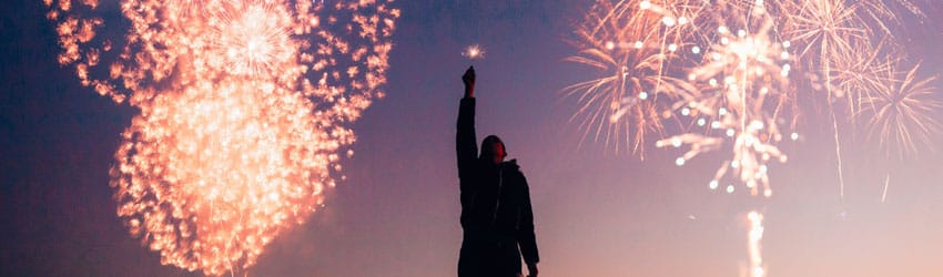 A celebrating person holds up sparklers in the sky where there are fireworks behind them.