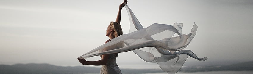 A blonde woman wearing a white gown holds up white gauzy fabric behind her, which appears to be blowing in the wind.