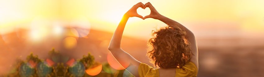 A woman makes a heart shape with her fingers with the sunset in the background.