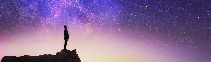 A silhouetted figure stands on a rock against the night sky, which is filled with blue, purple, and yellow hues. The white stars speckle the expansive sky.