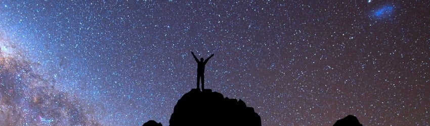 A shadowed figure stands on top of a cliff, facing the night sky.