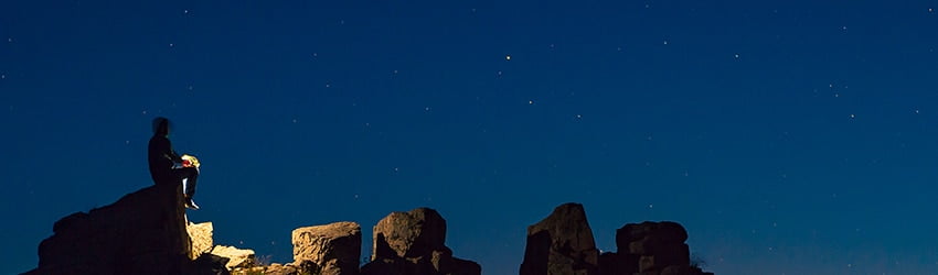 A man sits on a rock looking up at the stars, backlit by his gas lantern.