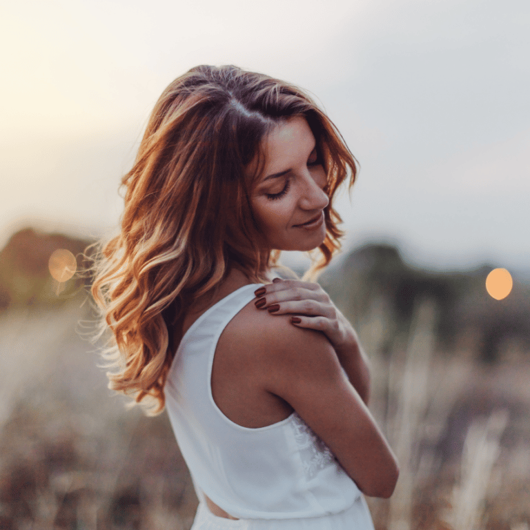 A woman embraces her shoulder, looking wistfully at the ground.