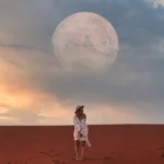 woman standing in the desert in white linen and a hat in front of a sky filled with a full moon