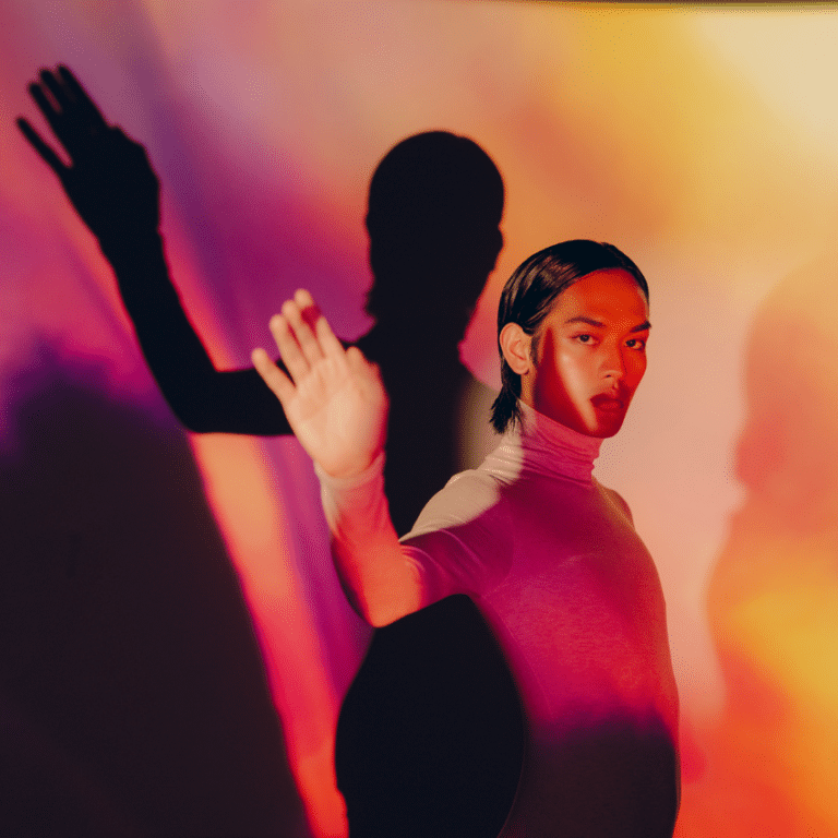 man in white turtleneck holding his hand up surrounded by orange and pink lighting