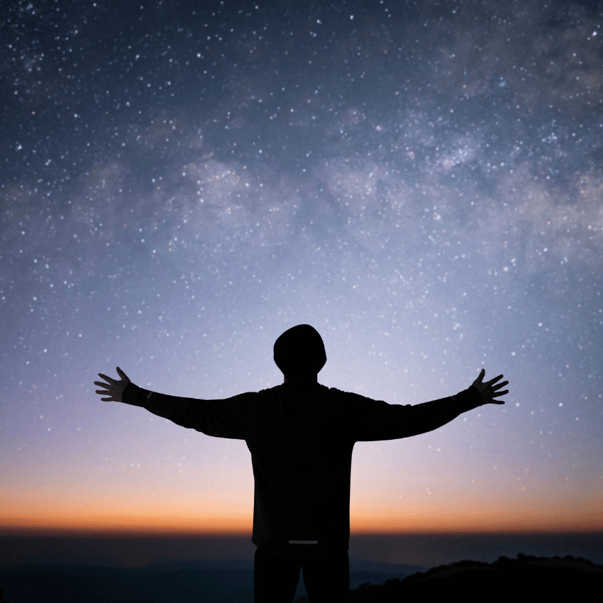 silhouette of a man with arms outstretched starring up at a blue starry sky