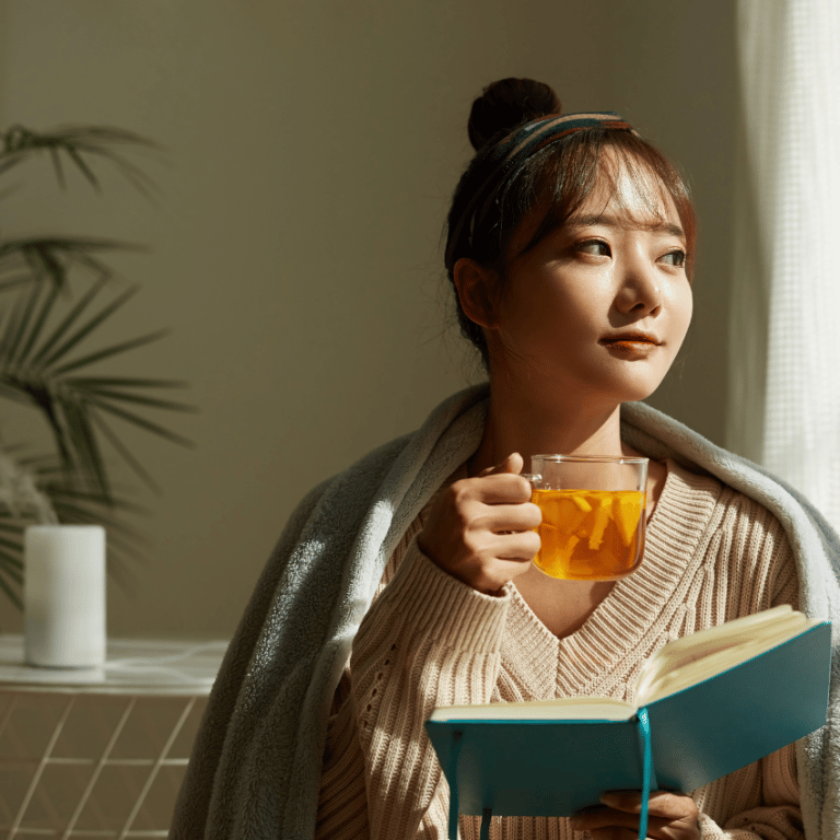 asian woman holding a cup of tea and a notebook gazing out the window