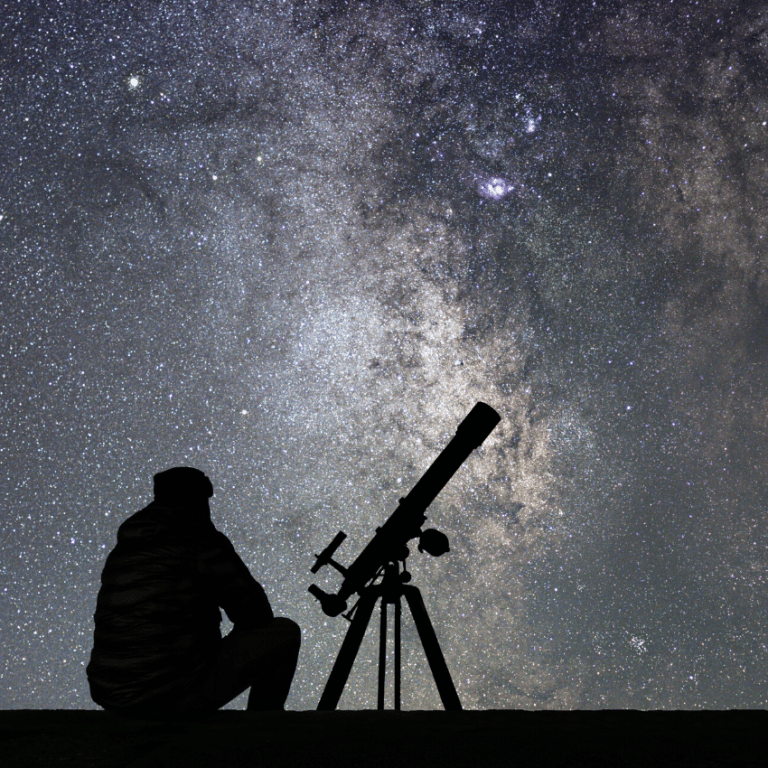 silhouette of a man sitting next to a telescope pointed up at a starry grey sky