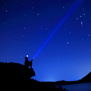 man sitting on the edge of a cliff shining a light up to a blue starry sky