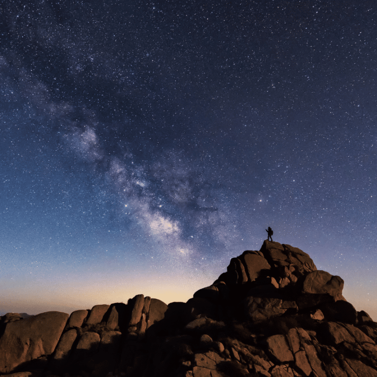 man at a distance standing on a clifftop with a starry blue and purple sky behind him