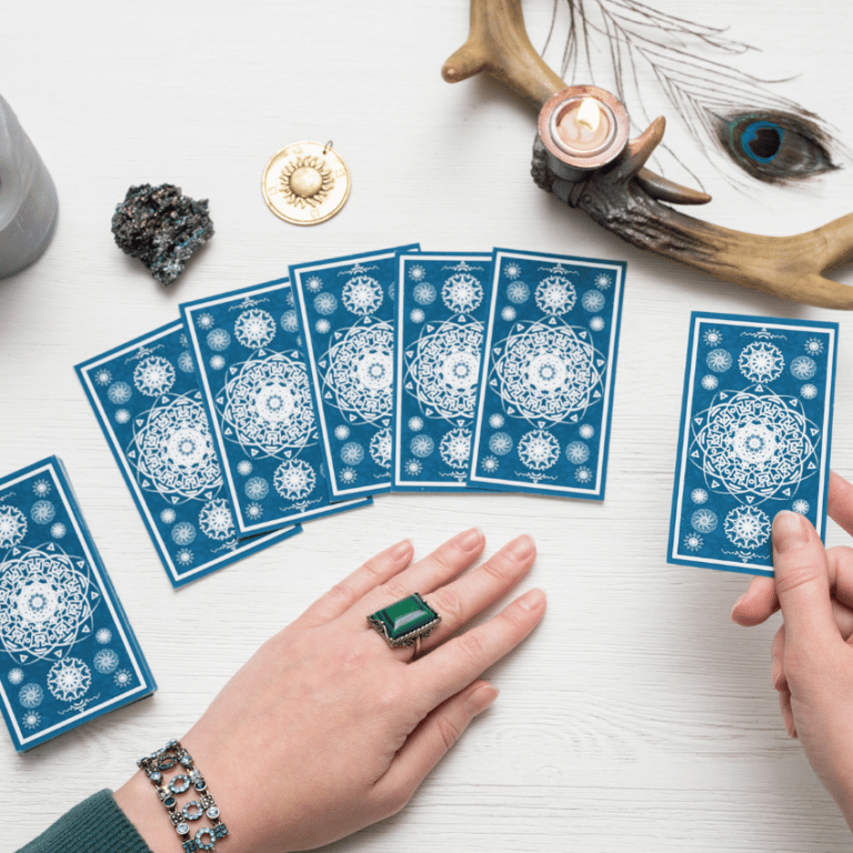 blue tarot card deck spread along a white table with a pair of hands holding one card with crystals and wood